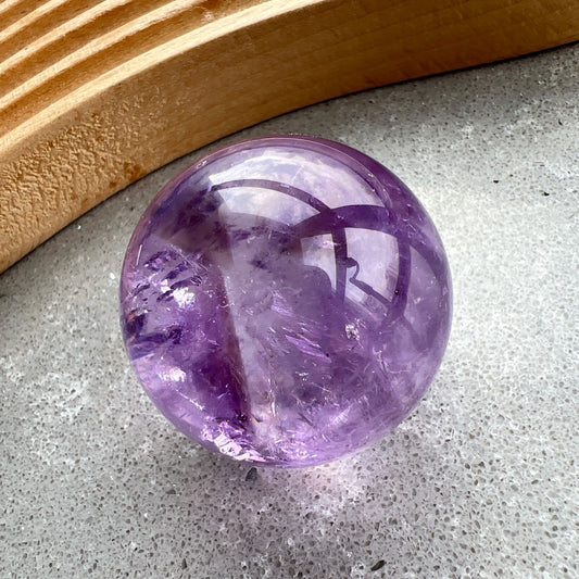紫水晶
【 圓潤之境，光之詩意 】｜ 彩虹光紫水晶球

如果說隨形是自然的率性，那麼水晶球便是大地最圓滿的溫柔。

這顆紫水晶球在純淨的紫色晶體中，包裹著歲月沈澱的天然冰裂紋與雲霧。 透過自然光線的折射，球體內部在不同角度下會閃耀出如夢似幻的彩虹光芒，彷彿將雨後的晴空濃縮於指尖。